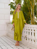 A woman in the Social Queen Feather-Trimmed Embellished Satin Caftan Dress stands by a white stone balustrade, wearing gold sandals and holding a woven handbag, with green plants in the background.