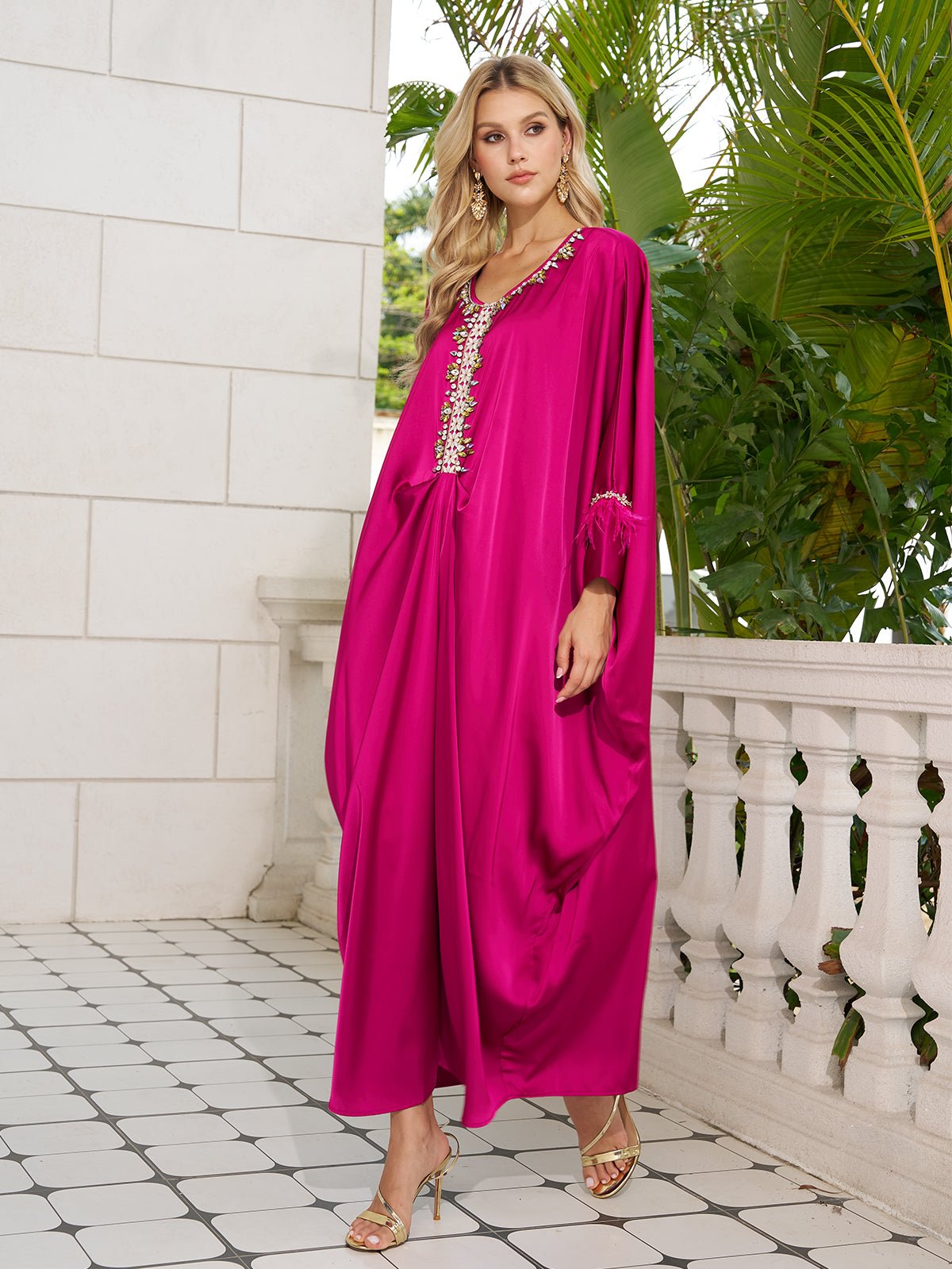 A woman stands on a tiled balcony in the Social Queen Feather-Trimmed Embellished Satin Caftan Dress, paired with gold heels. Lush greenery serves as the backdrop.