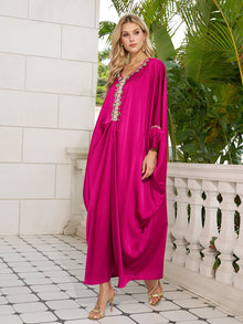 A woman stands on a tiled balcony in the Social Queen Feather-Trimmed Embellished Satin Caftan Dress, paired with gold heels. Lush greenery serves as the backdrop.