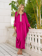 On a tiled terrace, a woman wears the Social Queen Feather-Trimmed Embellished Satin Caftan Dress in fuchsia, styled with gold sandals and a woven handbag. Lush green plants provide a vibrant backdrop.