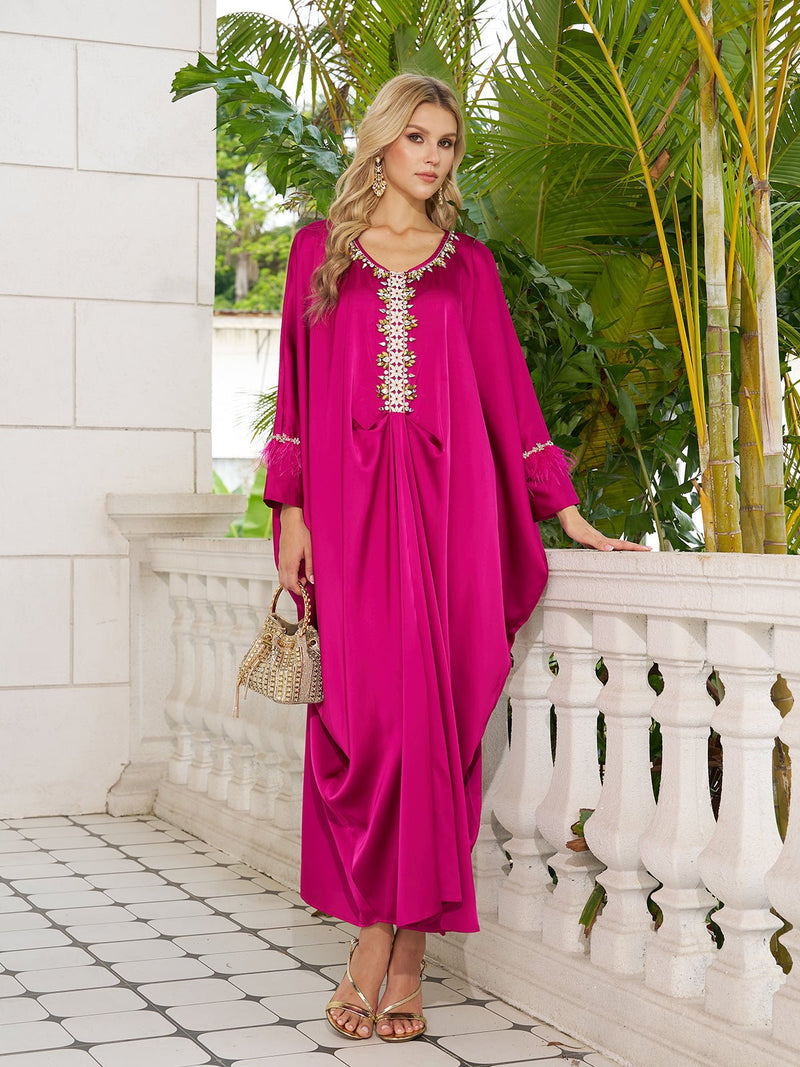 On a tiled terrace, a woman wears the Social Queen Feather-Trimmed Embellished Satin Caftan Dress in fuchsia, styled with gold sandals and a woven handbag. Lush green plants provide a vibrant backdrop.