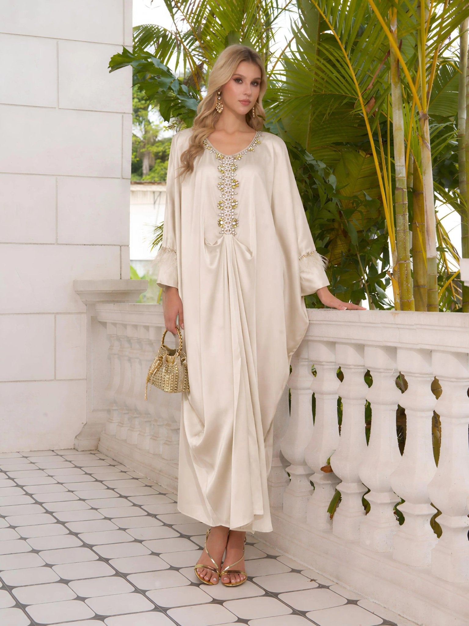 A woman in the Social Queen Feather-Trimmed Embellished Satin Caftan Dress stands on a tiled balcony with white columns and tropical plants, holding a textured handbag.