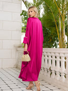 A woman in the Social Queen Feather-Trimmed Embellished Satin Caftan Dress and heels stands on a tiled balcony, holding a woven handbag, with greenery and a white stone wall behind her.