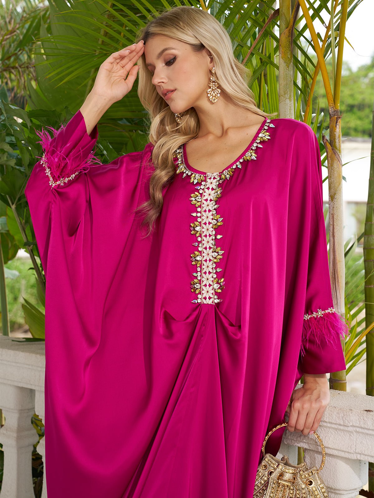 A woman in the Social Queen Feather-Trimmed Embellished Satin Caftan Dress with gold earrings stands near green plants, holding a gold handbag and touching her forehead.