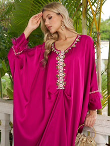 A woman in the Social Queen Feather-Trimmed Embellished Satin Caftan Dress with gold earrings stands near green plants, holding a gold handbag and touching her forehead.