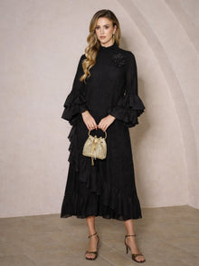 A woman stands indoors in a Social Queen Floral Appliqué Embroidered Ruffle Dress, holding a small gold handbag and wearing high-heeled sandals.