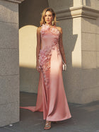 A woman stands outside in the Social Queen Floral Appliqué Satin Fishtail Gown, styled with a clutch, gold heels, and earrings against a stone building.