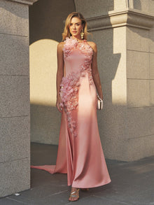 A woman stands outside in the Social Queen Floral Appliqué Satin Fishtail Gown, styled with a clutch, gold heels, and earrings against a stone building.