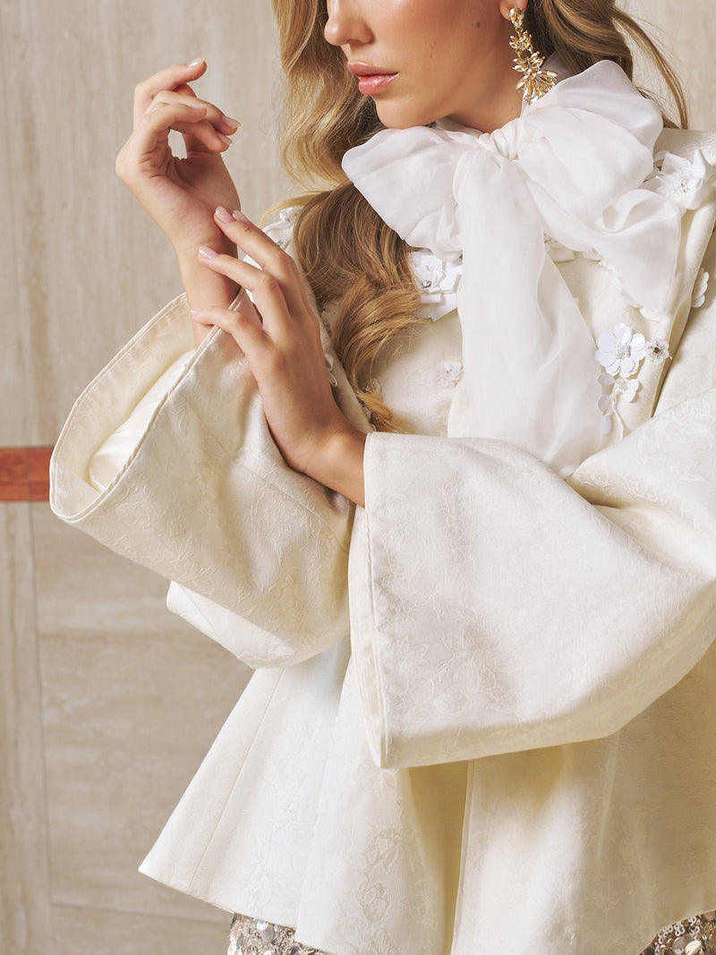 A woman models the Social Queen Floral Appliqué Satin Jacket—cream with wide sleeves, hand-sewn floral details, and a white neck bow—paired with statement earrings against a beige background.