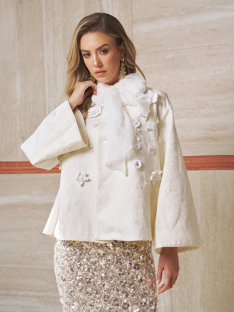 A woman in the Social Queen Floral Appliqué Satin Jacket stands against a beige marble wall, featuring hand-sewn florals, a textured cream finish, and a statement white bow at the neck.