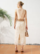 A woman stands on terracotta tiles facing away, wearing the Social Queen Floral Appliqué Sheer Sleeveless Top, a textured white skirt with fringe, heeled sandals, and holding a small woven handbag.