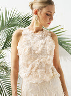 A woman models the Social Queen Floral Appliqué Sheer Sleeveless Top with 3D floral details, paired with large tassel earrings, standing before green palm leaves.