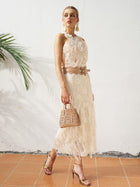 A woman stands on a tiled floor against a white wall with a palm leaf, wearing the Social Queen Floral Appliqué Sheer Sleeveless Top and holding a small beige handbag, paired with gold heels.