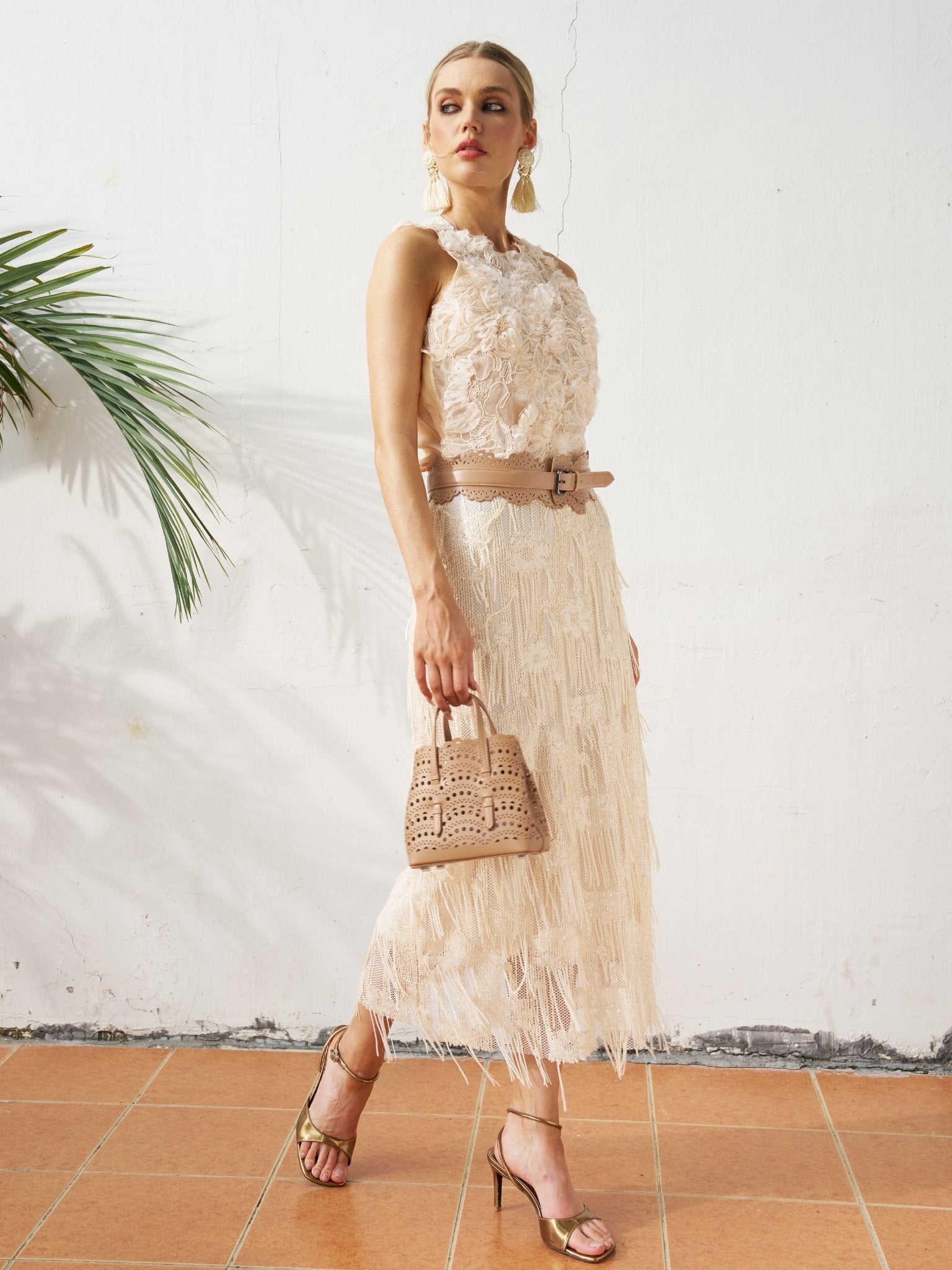 A woman stands on a tiled floor against a white wall with a palm leaf, wearing the Social Queen Floral Appliqué Sheer Sleeveless Top and holding a small beige handbag, paired with gold heels.