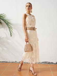 A woman stands on a tiled floor against a white wall with a palm leaf, wearing the Social Queen Floral Appliqué Sheer Sleeveless Top and holding a small beige handbag, paired with gold heels.