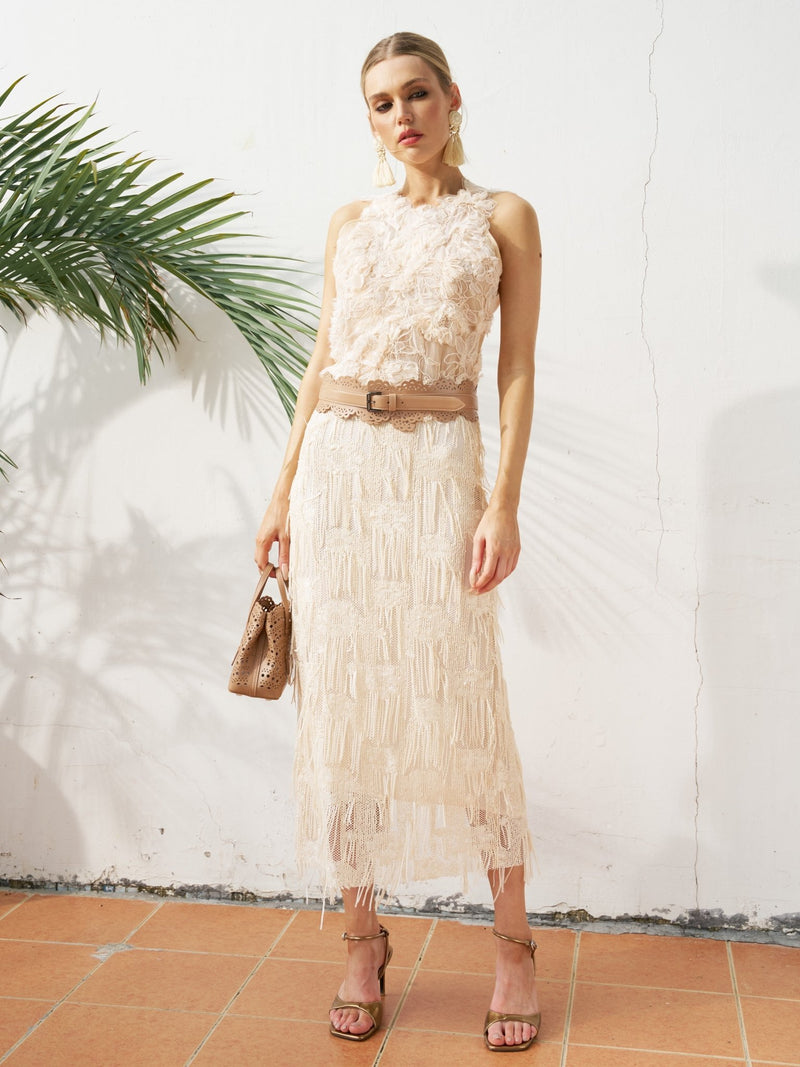 A woman in the Social Queen Floral Appliqué Sheer Sleeveless Top stands near a white wall and green plant, holding a matching purse and wearing the top with a brown belt on terracotta tiles.