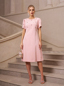 A woman stands on stone stairs wearing the Floral Beaded Puff Sleeve Dress by Social Queen, a light pink, knee-length hand-beaded floral dress with short sleeves—an elegant choice for a wedding guest.