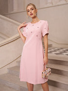 A woman stands on stairs in an elegant setting, wearing the Social Queen Floral Beaded Puff Sleeve Dress in light pink with floral embellishments, and holding a small matching floral purse.