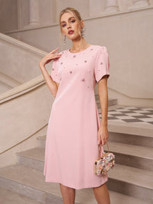 A woman stands on stairs in an elegant setting, wearing the Social Queen Floral Beaded Puff Sleeve Dress in light pink with floral embellishments, and holding a small matching floral purse.