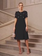 A woman stands on a staircase wearing the Social Queen Floral Beaded Puff Sleeve Dress, paired with silver sandals and holding a small decorative handbag.