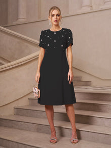 A woman stands on a staircase wearing the Social Queen Floral Beaded Puff Sleeve Dress, paired with silver sandals and holding a small decorative handbag.