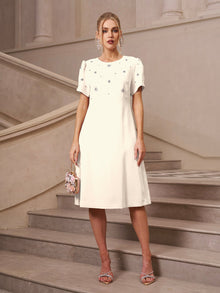 A woman stands on a staircase wearing the Social Queen Floral Beaded Puff Sleeve Dress, holding a small handbag and sporting open-toe heels.