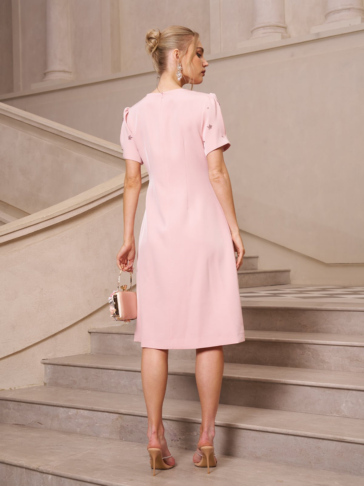 A woman in the Social Queen Floral Beaded Puff Sleeve Dress and heeled sandals stands on a staircase, facing away and holding a small matching handbag.