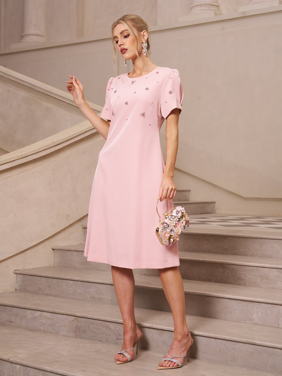 A woman stands on stairs wearing the Social Queen Floral Beaded Puff Sleeve Dress, paired with silver heels and a floral purse—an ideal choice for a wedding guest.