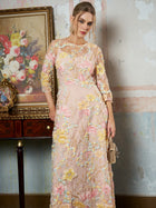A woman indoors stands by a table with framed art and flowers, holding a small gold handbag. She is wearing the Social Queen Floral Beaded Sheer Gown—a long, pastel A-line evening dress with 3D floral appliqué.