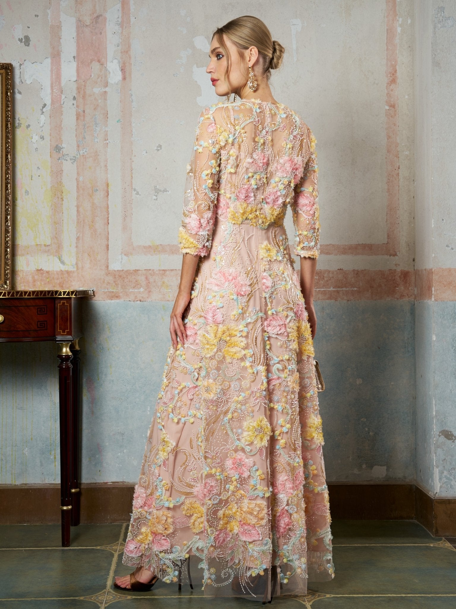 A woman stands indoors with her back to the camera, wearing the Social Queen Floral Beaded Sheer Gown featuring pink, yellow, and green floral embroidery and delicate 3D appliqué, looking to the left.