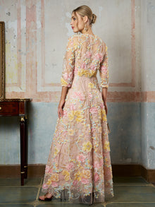 A woman stands indoors with her back to the camera, wearing the Social Queen Floral Beaded Sheer Gown featuring pink, yellow, and green floral embroidery and delicate 3D appliqué, looking to the left.