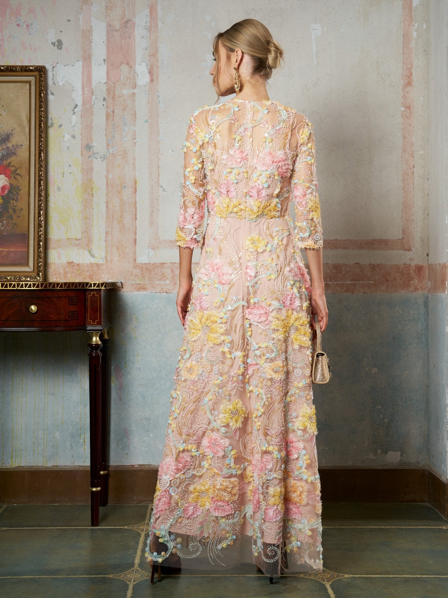 A woman stands indoors facing away, wearing the Social Queen Floral Beaded Sheer Gown with long sleeves, 3D floral appliqué, and holding a small clutch.