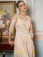 A woman in the Social Queen Floral Beaded Sheer Gown stands indoors beside a framed floral painting on a wooden table.