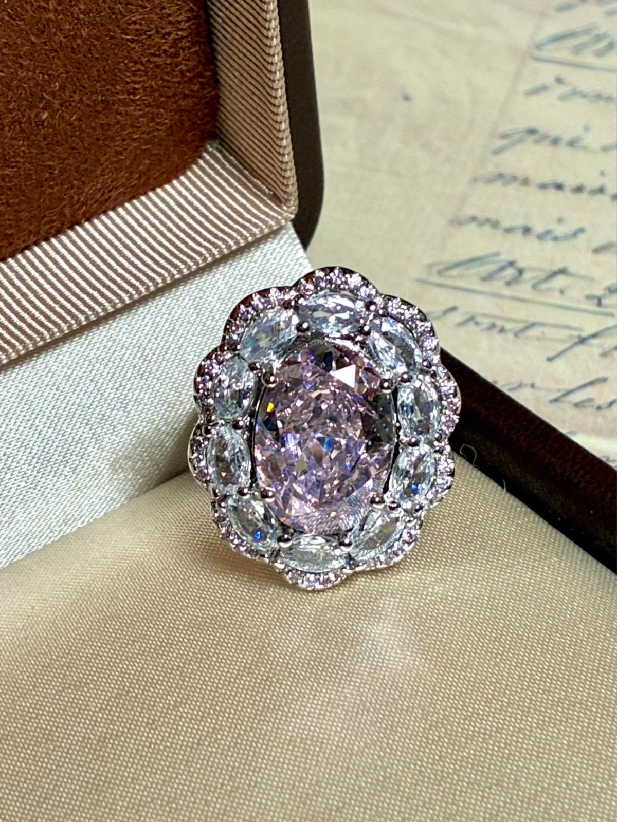The Social Queen Floral Crystal Ring features a large oval pink gemstone surrounded by sparkling clear stones, beautifully presented in a jewelry box—perfect for adding glamour to any outfit.