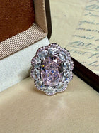 The Social Queen Floral Crystal Ring features a large oval pink gemstone surrounded by sparkling clear stones, beautifully presented in a jewelry box—perfect for adding glamour to any outfit.