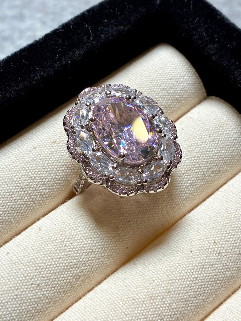 The Social Queen Floral Crystal Ring, featuring a large oval pale pink gemstone encircled by clear stones, is presented in a cream-colored ring box with a black edge.