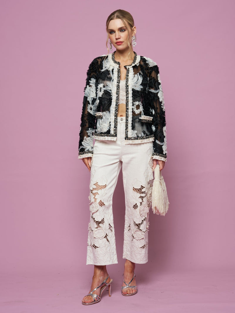 A woman stands against a pink background wearing Social Queen’s Floral Cutout High-Rise Jean, a black and white jacket with floral cutout embroidery, silver heels, and holding a beaded fringe bag.