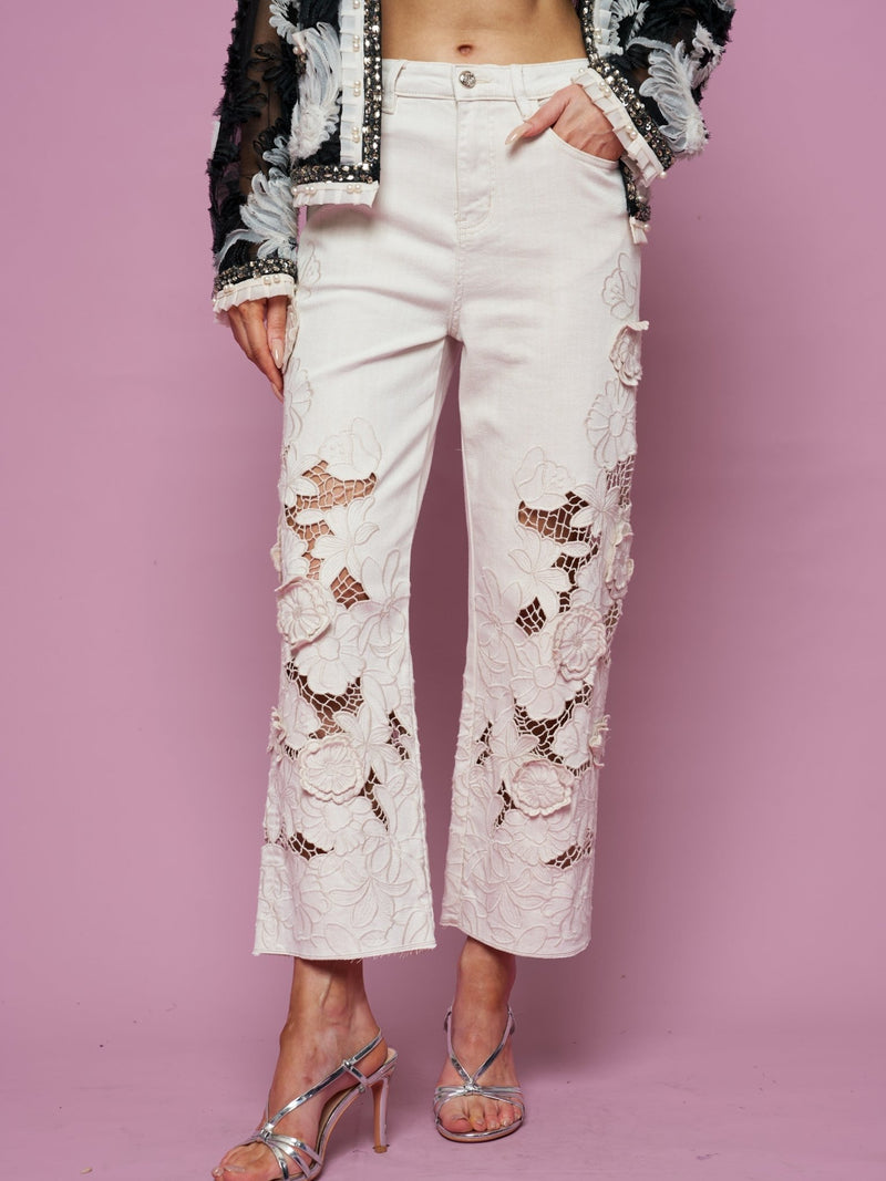 A person in Social Queen’s Floral Cutout High-Rise Jean, a black and white textured jacket, and silver high-heeled sandals stands against a pink backdrop.