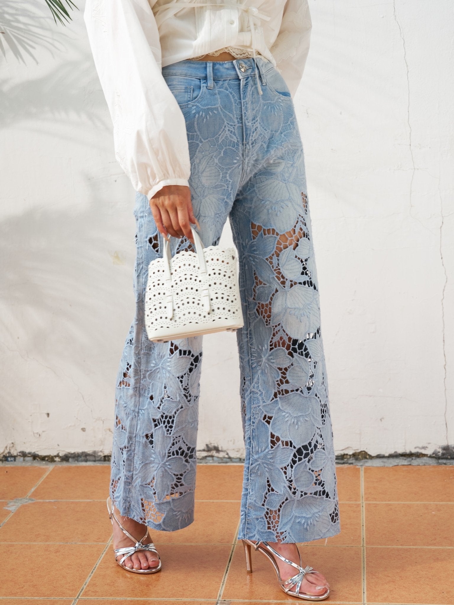 A person wears Social Queen Floral Cutout Stretch Denim Jean in light blue with silver heels, holding a small white woven handbag, standing on a tiled floor against a white wall.