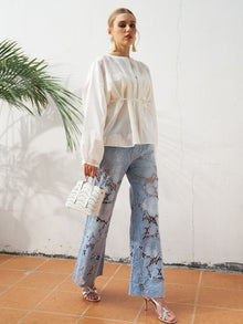 Indoors, a person wears a loose white blouse, Social Queen Floral Cutout Stretch Denim Jean, silver heels, and holds a white handbag. A palm leaf appears on the left.