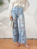 A person stands on tiled flooring wearing Social Queen's Floral Cutout Stretch Denim Jean in light blue, styled with a white long-sleeve blouse, silver sandals, and a small white handbag.