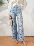 A person stands on tiled flooring wearing Social Queen's Floral Cutout Stretch Denim Jean in light blue, styled with a white long-sleeve blouse, silver sandals, and a small white handbag.