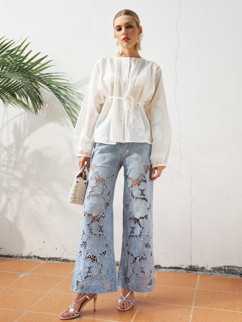 A woman indoors wears a white long-sleeve blouse, Social Queen Floral Cutout Stretch Denim Jean in light blue, silver heels, and holds a white handbag; a palm leaf appears in the background.