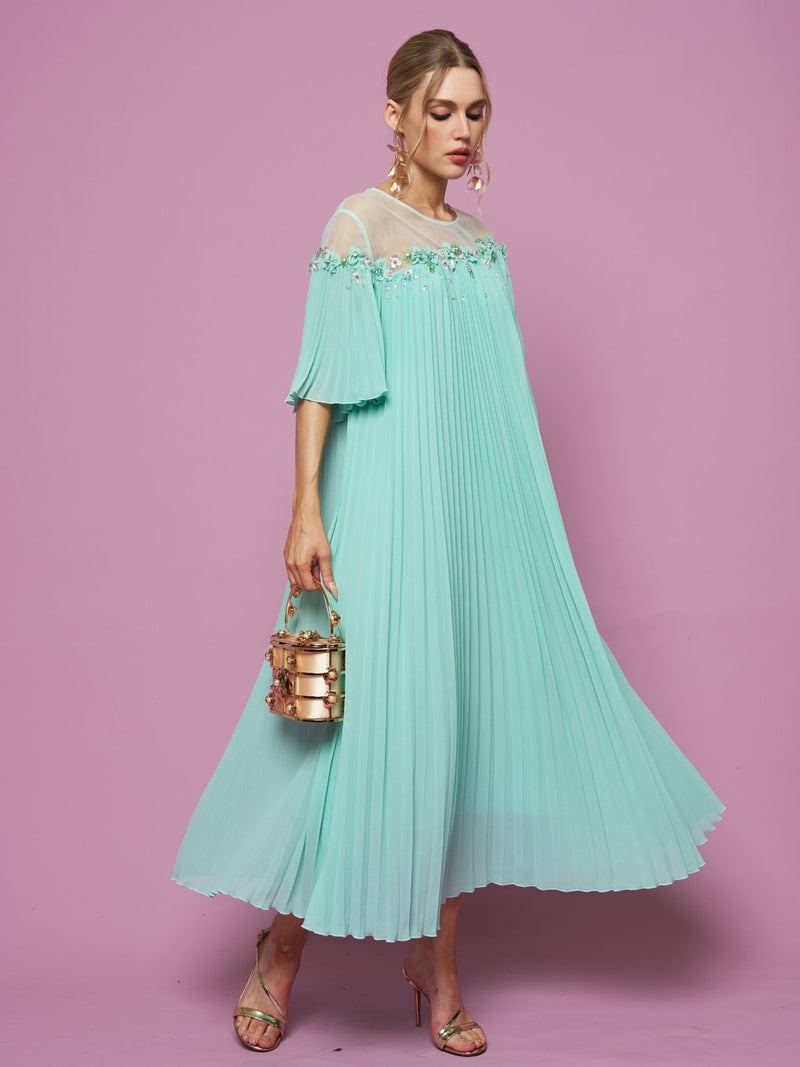 A woman in the Social Queen Floral Embellishment Pleated Dress with a relaxed silhouette holds a metallic handbag, standing against a pink background.