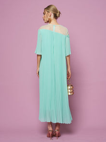 A woman faces away against a pink background, wearing the Social Queen Floral Embellishment Pleated Dress in light blue with a relaxed silhouette, paired with pink heels and a small gold and white handbag.