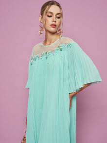 A woman poses against a pink background wearing the Social Queen Floral Embellishment Pleated Dress, styled with statement earrings and a small purse.