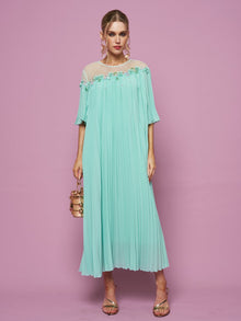 A woman stands against a pink background wearing the Social Queen Floral Embellishment Pleated Dress, paired with a small gold handbag and strappy heels.