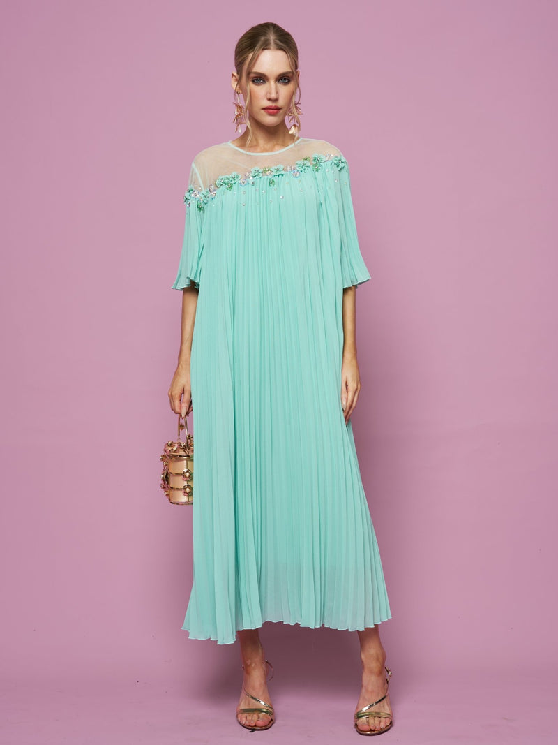 A woman stands against a pink background wearing the Social Queen Floral Embellishment Pleated Dress, paired with a small gold handbag and strappy heels.