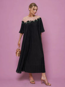 A woman in Social Queen's Floral Embellishment Pleated Dress stands against a pink background, pairing the black beaded mesh dress with a small handbag and gold heels.