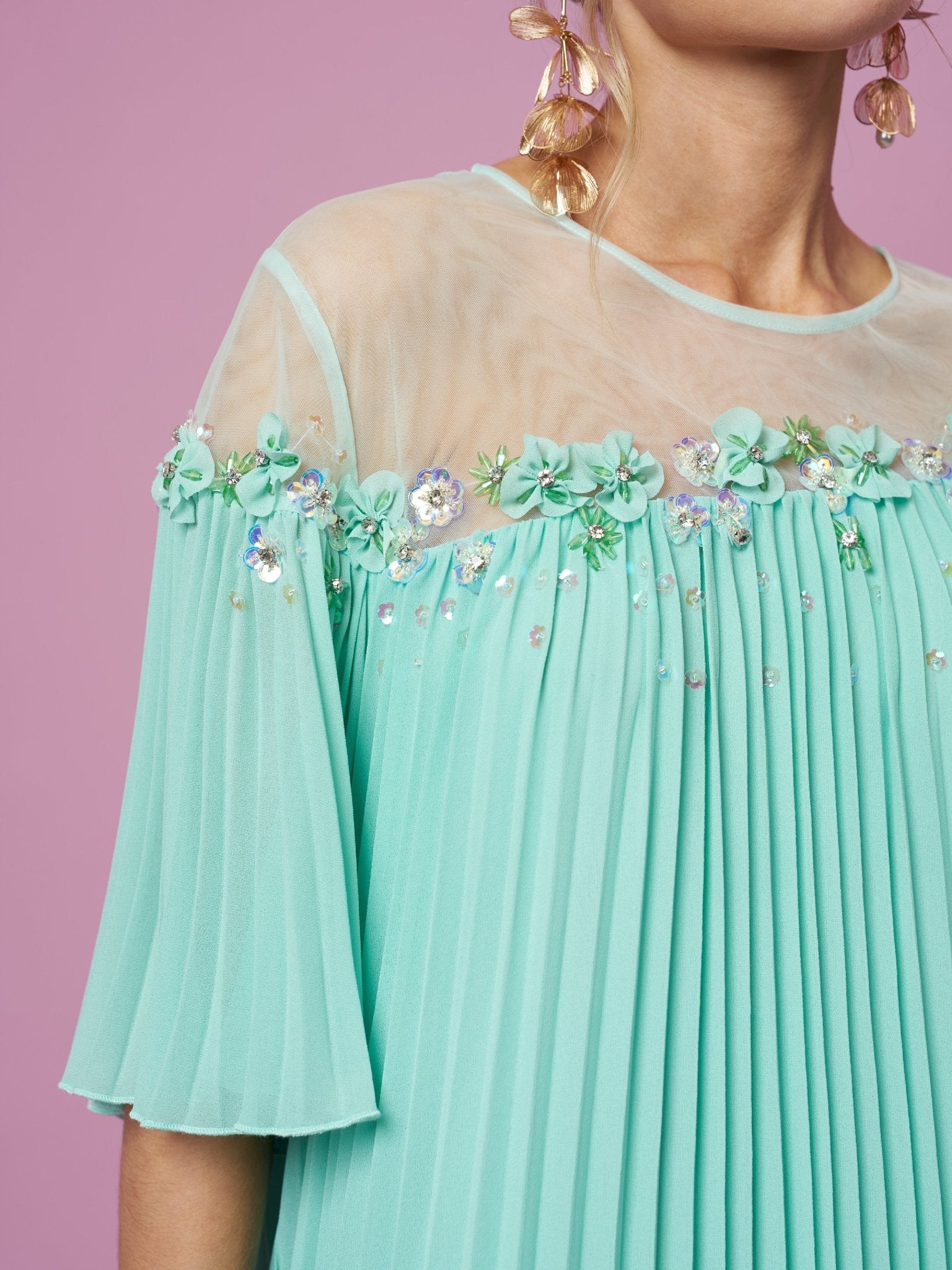 A person stands against a pink background wearing the Social Queen Floral Embellishment Pleated Dress in mint green, featuring sheer panels, floral beadwork, sequins, and large gold leaf-shaped earrings.